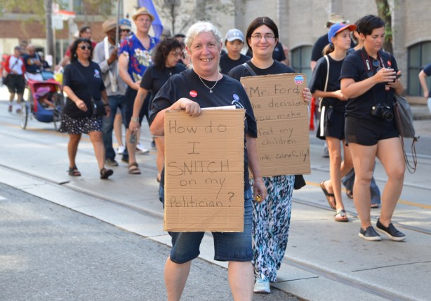 labour Day parade on Queen St West, two women carry brown cardboard signs, one says How do I snitch on my politician? and the other talks about policies affecting education ofchildren 