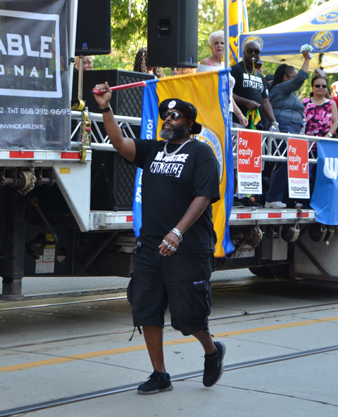 labour Day parade on Queen St West, a black man waves a union flag, wearing a black t shirt with the words No Justice No Peace. Some other people are riding on a flat bed truck behind him, with posters that say Pay Equality for all 