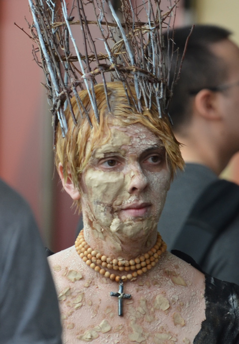a woman wearing a tall crown made of sticks, three strands of yellow beads and a cross around her neck, lots of make up to make her face look mottled and bumpy