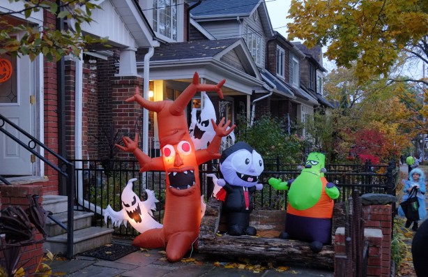 Halloween night, early evening, not quite dark, a street scene where one house has a lot of inflatable characters on the lawn as decorations