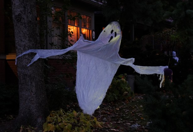 ghost made of white fabric waving in the breeze in front of a house, night time, window lights on 