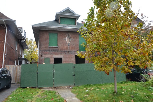 large square brick house from the early 1900s, windows boarded up and green plywood hoardings in front