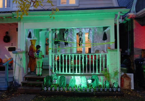 Halloween night, trick or treaters at the door of a house with lots of tombstones and skulls, also the porch is lit by an eerie green light. 
