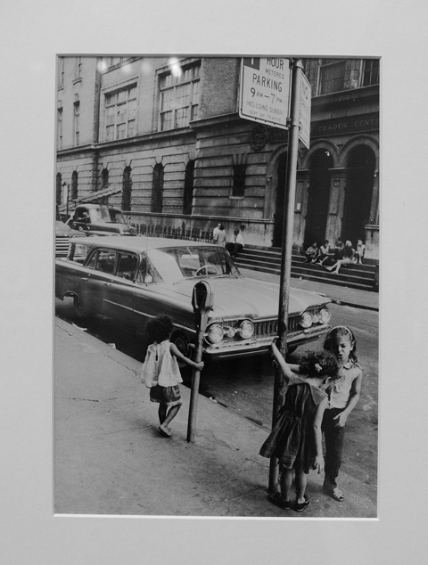 old photo from the 1960s, 3 children play on the sidewalk by a car that is parked on the street