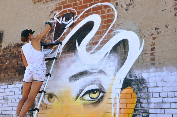 Anya painting a mural of a woman with wavy white and black hair, yellow face, yellow eyes, she's standing on a ladder