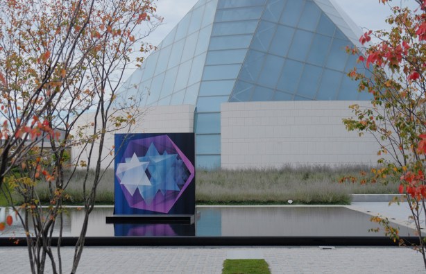 A painting in blue, pink, and purple, of stars, by Jacid Jah, in a reflecting pool with the Ismaili Centre behind it 