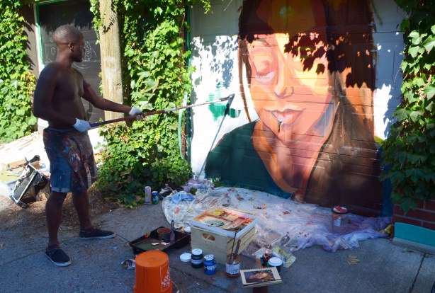 using a long handled brush to paint a mural of a woman's face on a garage door 