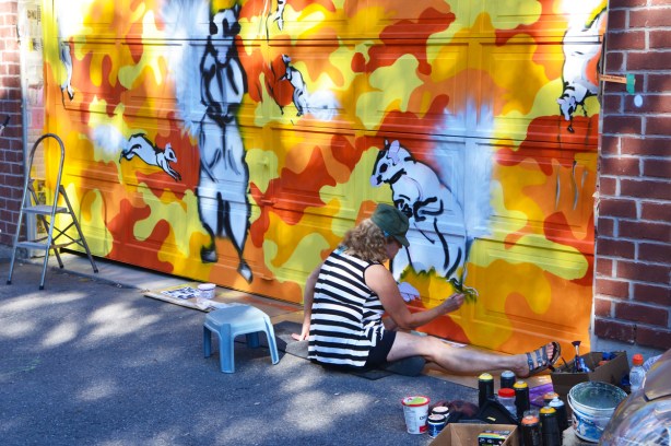 artist sitting on the ground while painting a mural on a garage door in a lane 
