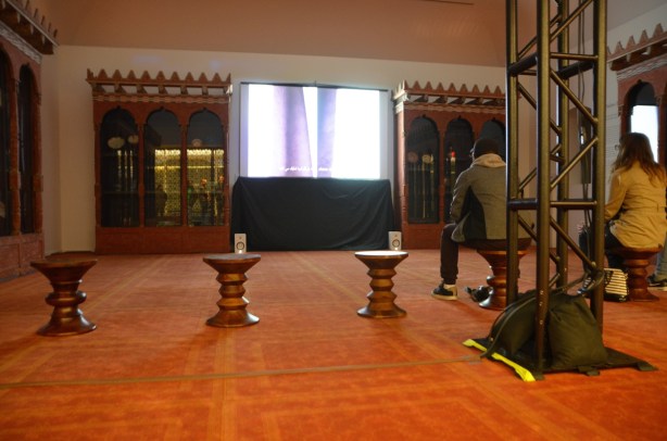 a room in the Aga Khan that is showing a video made in Tehran, two people are sitting on stools as they watch it, orange carpet on the floor, old decorated bookcases against the walls.