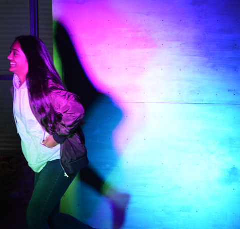 woman runs past a white screen that is being lit with coloured lights, making her shadow coloured