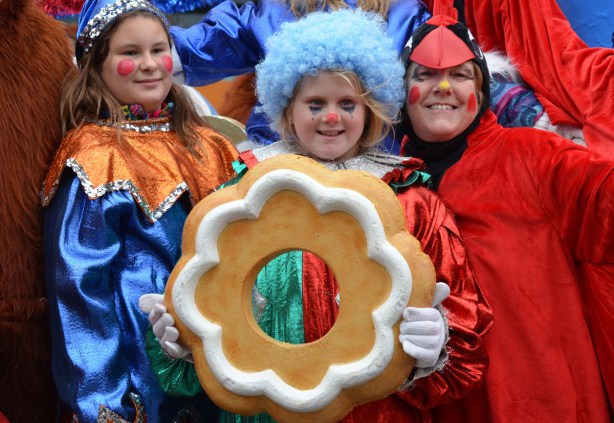 three people involved with the Santa Claus parade pose for a picture. A woman in a red cardinal costume, a girl with a light blue wig holding an oversized gingerbread cookie with white icing, and another girl in a shiny blue and orange outfit 