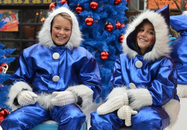 two girls on a float at the Santa Claus parade, dressed in shiny blue clothes with fuzzy white furry trim, smiling and looking at the camera 