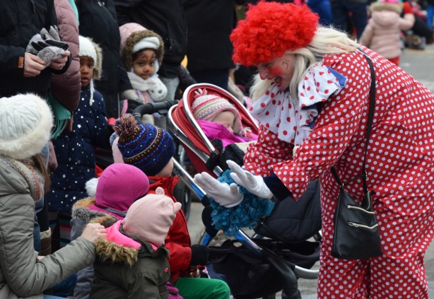 a clown in polka dot outfit and red wig bends down to high five with kids on the sidewalk who are watching the parade 