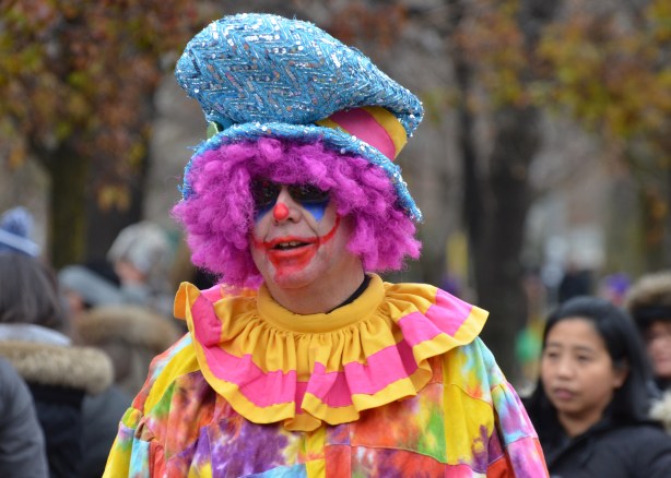 a male clown in pink wig and pink and yellow ruffle collar, large floppy blue hat and lots of make-up on his face 