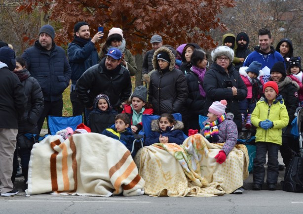 a group of people on the sidewalk watching a parade, kids in front on chairs and under blankets, parents behind standing. 