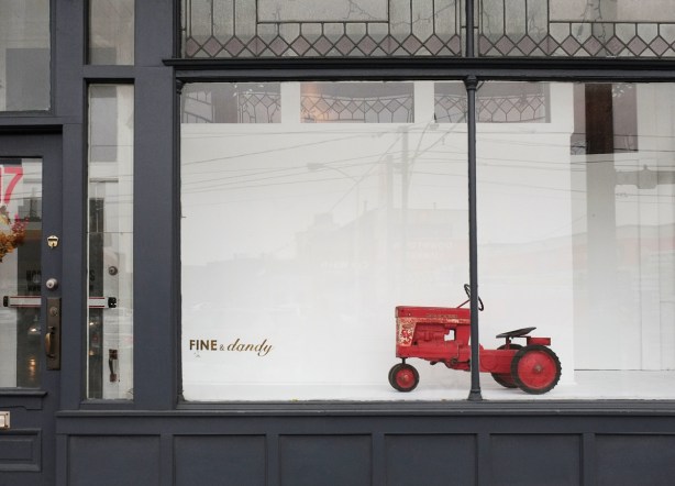a red toy tractor, old fashioned, in the window of fine and dandy on Dundas Street, white back drop behind the tractor, the building is dark grey 