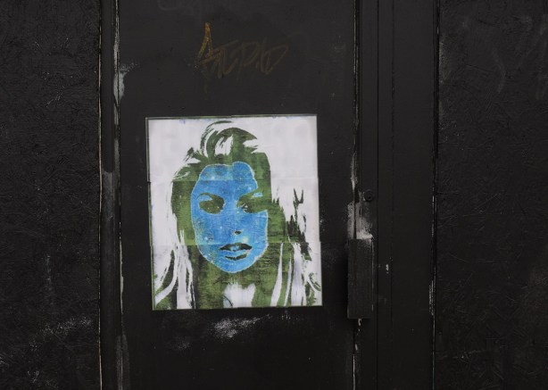 a paper paste up of woman's face in green and blue (green skin and blue hair) on a very black wall and door 