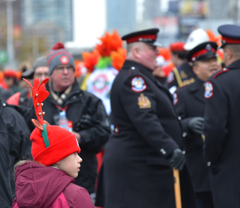 a girl with a red hat with red reindeer ears stands on the street on a parade route, a group of policemen is in the background. 