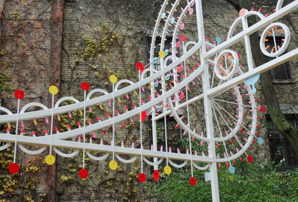 part of a white metal sculpture of a treble clef and a line of music, with yellow and red lights, in a garden, in a city with a brick wall behind it 