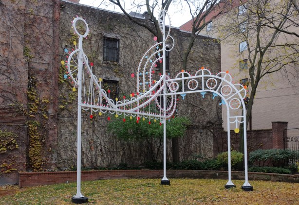  a white metal sculpture of a treble clef and a line of music, with yellow and red lights, in a garden, in a city with a brick wall behind it 