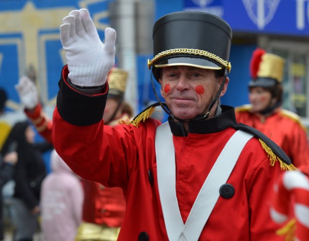 a man dressed as a tin soldier walks in a parade, waving to the crowd wearing white gloves & red uniform