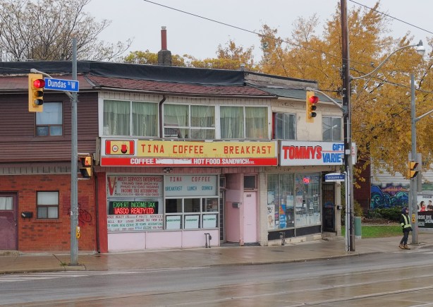 restaurant and store, rainy day, wet sidewalk and street in front of it, Tina Coffee and Breakfast restaurant, and Tommys Gift & Variety, pink door between the two, two storeys, lots of windows in the storey above Tina's. 