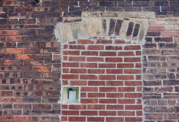 a brick wall, an old arched window has been bricked in, leaving a small window, in the window is a roll of toilet paper 