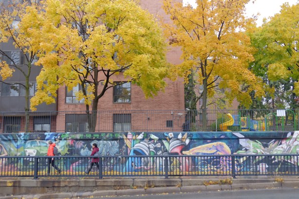 people walking on a sidewalk that passes by a long mural painted by wallnoize, many small murals joined together, apartment buildings with large trees with yellow autumn leaves in the background, Bloor West, 