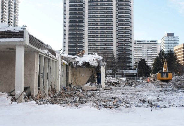 small part of a school remains, debris scattered on the snow, digger at work in the background, apartment building in the distance