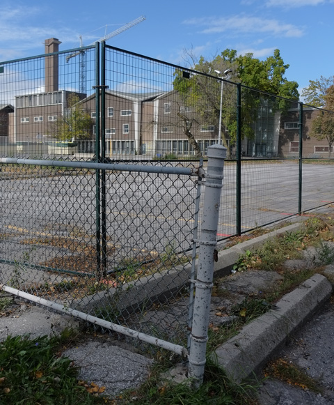 crooked chainlink fence posts at the corner of a schoolyard, with metal construction fence inside that, school in the background, large paved area in front of the school 