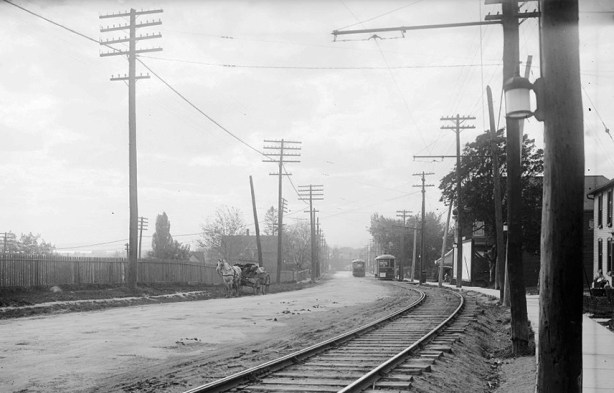 old black and white picture from 1922 of a dirt street with a street car track, hydro poles beside the road and a house 