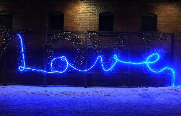 a band of blue light writes the word love in cursive, laid on top of a LOVE sign that is covered with locks. 