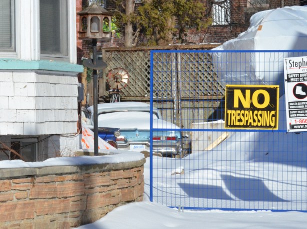 a no trespassing sign on a wire fence, snow covered driveay, two old cars parked in the backyard, beyond the fence 