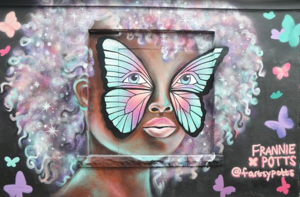 painting by Frannie Potts, a street art face on a garage, woman, with large curly afro style hair in pale colours, with a butterfly painted covering her eyes and cheeks. 