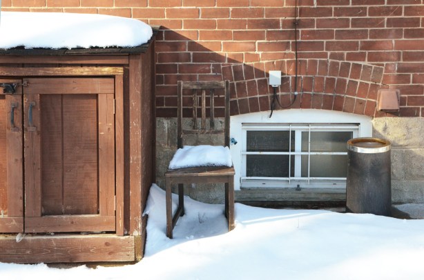 wooden chair outside, against the side of a house, snow on it. 