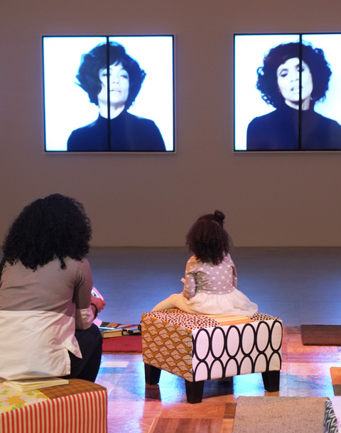 a mother and young daughter sit on seats in a room at the Art Gallery of Ontario, watching videos by Mickalene THomas as part of her Femmes Noires exhibit