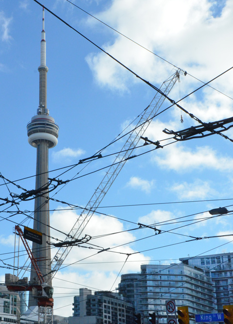 CN tower, blue sky with some clouds, a construction crane and lots of streetcar wires. There are some glass condo buildings in the background. Street sign that says King St., traffic lights