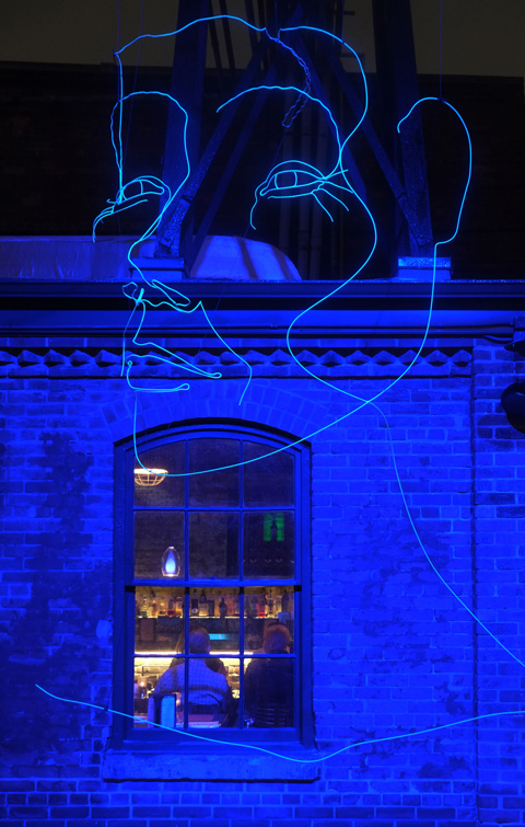 blue lights shine against an exterior brick wall of a restaurant, window - looking in the window you can seen people sitting at a table. A large art installation of a woman's face outline in blue light hovers above the window. 