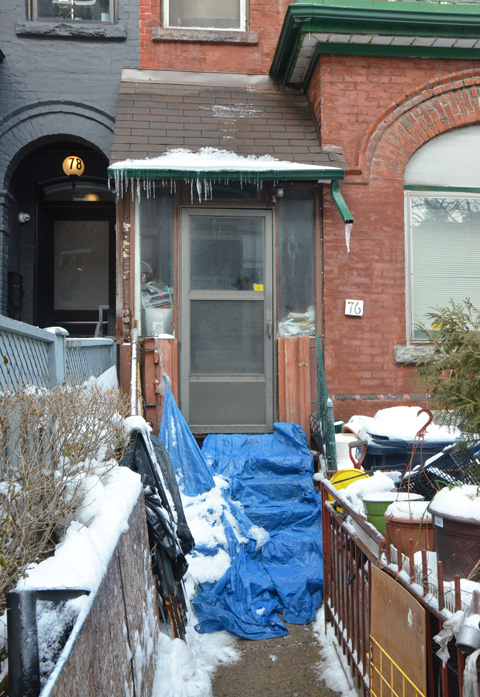 a blue tarp covers the front steps of a brick house, small fence between the front lawn and walkway, lots of junk in the front yard.