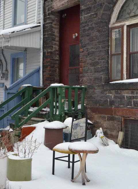 greyish stone house with dark red front entrance, small porch with dark green wood railing, table and chair left on front yard, covered with snow