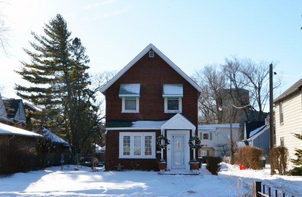 a typical old Toronto two storey house with peaked roof, reddish brick, two wondows upstairs, one large window downstairs, white front door with a small roof over the door, lots of yard 