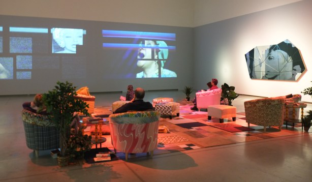 people sitting, furnishings, comfy armchairs and a carpet on the floor, turing a room at the AGO into a livingroom, one wall has video (or videos) playing over its entire surface