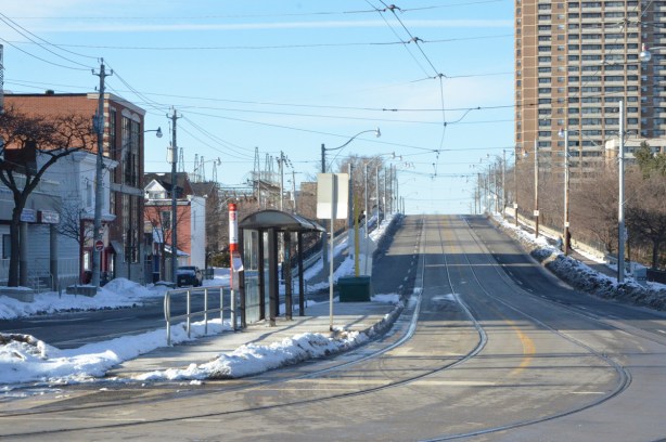 looking north up Main street from Gerard, streetcar tracks with a bus shelter in the middle of the street. old style bus shelter, Main street then goes up, as a bridge over the train tracks. Highrise apartment building in the background. 