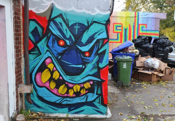 gnarly monster face painted on the side of a small wall, bllue, with big yellow teeth and small red eyes 