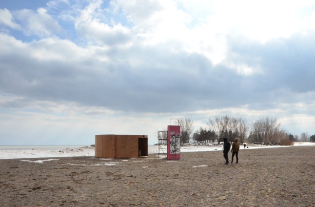 the circular art installation 'Mind Station', wood, on the beach beside Lake Ontario on a winter day, two men walking towards it, trees in the distance 