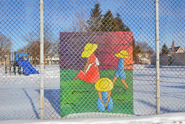 colourful painting behind a chainlink fence in a school yard, winter, snow on the ground around it, picture is of three kids in large yellow hats, playing on green grass 