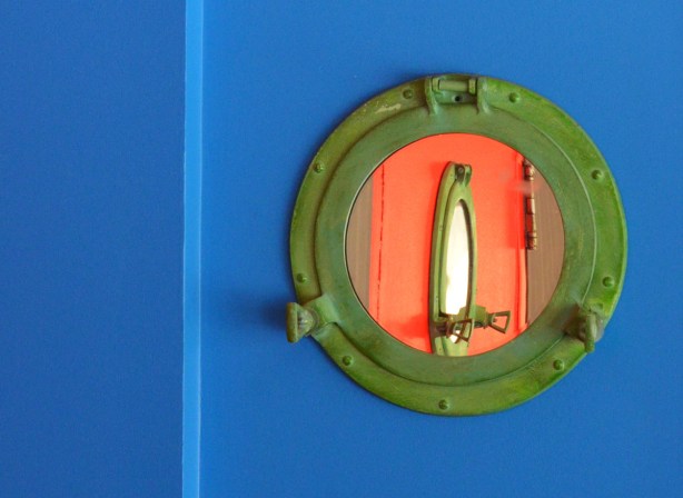 a mirror shaped like a porthole with a green frame, on a bright blue wall, reflection of another porthole but on an orange wall in the mirror