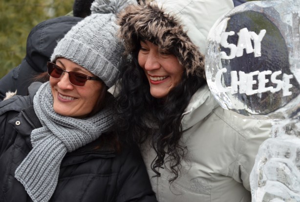 two women pose beside an ice sculpture that says Say Cheese. One woman is wearing a grey toque and scarf and the other woman is wearing a white parka with a fur lined hood