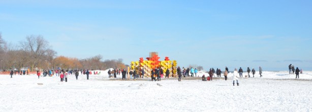 panorama of Woodbine Beach on a February day, art installation in yellows and oranges, lots of people, snow on the ground, winter clothes, Lake Ontario, blue sky with one wispy cloud