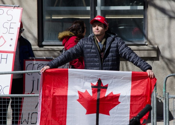 a young man wearing a red MAGA baseball cap holds a Canadian flag on which a black cross has been drawn 